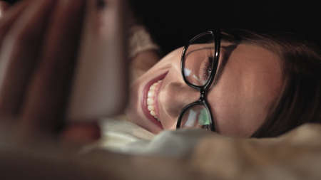 Woman with glasses looks at smart phone and lies on bed, smile on faceの写真素材