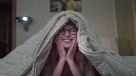 woman rejoices watching comedy at home, girl lying on the bed under the blanketの写真素材