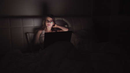 woman in glasses works on a laptop in the dark. portrait of a girl spending time at the computer late in the eveningの写真素材
