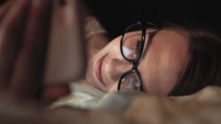 Woman with glasses looks at smart phone and lies on bed, smile on faceの写真素材