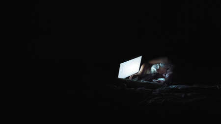 Nice girl watching something on a laptop monitor. Night atmosphere in the apartment. Laptop screen illuminates the girls faceの写真素材