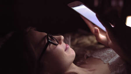 Side view of a happy teen browsing and watching media in a smart phone lying on a bed in the night at homeの写真素材