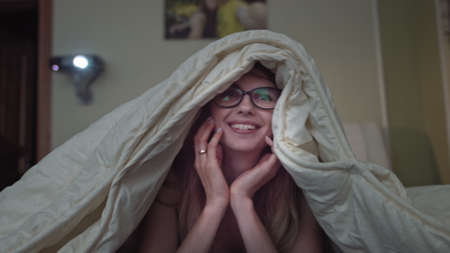 woman rejoices watching comedy at home, girl lying on the bed under the blanketの写真素材