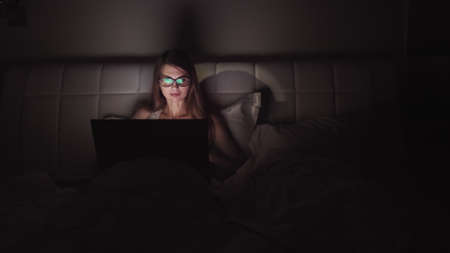 woman in glasses works on a laptop in the dark. portrait of a girl spending time at the computer late in the eveningの写真素材