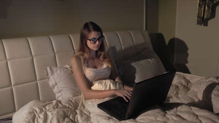 Young good looking girl in glasses typing and working on the laptop computer as freelancer while sittng at home in the cozy dark living room with a cat. At night. Stay homeの写真素材
