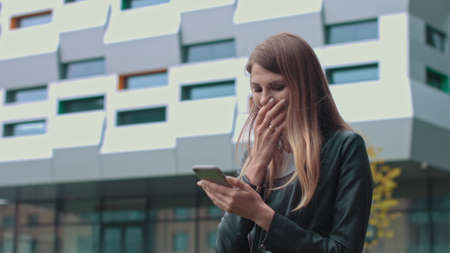 Young Attractive Girl with Long Brunette Hair walking in the Street, Having Mobile Phone in her Hands. Typing Messages and Smiling while sending. Having fashionable clothes.の写真素材