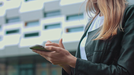 Young attractive girl in stylish clothes on the background of a modern building, looks at the phone...