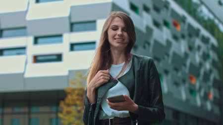 Good looking girl with Long Brown Hair Looking at Her phone Typing and Smiling. Enormous industrial Building at the Background. Green Bushes and Trees. Smart clothes. Natural makeup.の写真素材