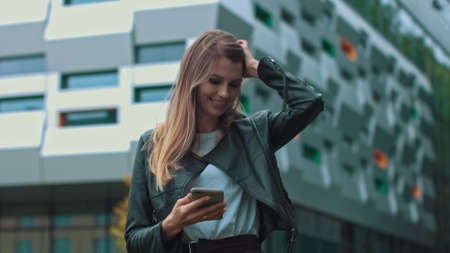 Good looking girl with Long Brown Hair Looking at Her phone Typing and Smiling. Enormous industrial Building at the Background. Green Bushes and Trees. Smart clothes. Natural makeup.の写真素材