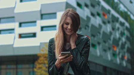 Good looking girl with Long Brown Hair Looking at Her phone Typing and Smiling. Enormous industrial Building at the Background. Green Bushes and Trees. Smart clothes. Natural makeup.の写真素材