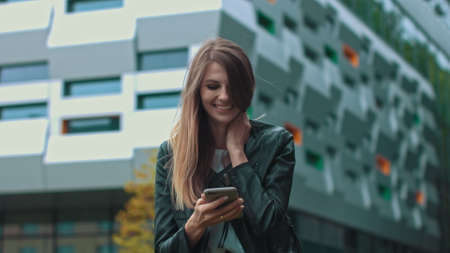 Good looking girl with Long Brown Hair Looking at Her phone Typing and Smiling. Enormous industrial Building at the Background. Green Bushes and Trees. Smart clothes. Natural makeup.の写真素材
