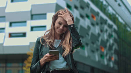 Good looking girl with Long Brown Hair Looking at Her phone Typing and Smiling. Enormous industrial Building at the Background. Green Bushes and Trees. Smart clothes. Natural makeup.の写真素材