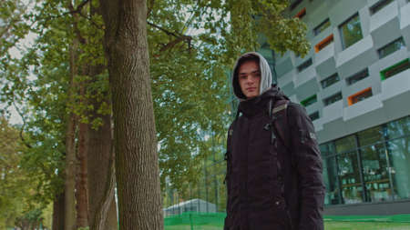 Portrait of a hipster young man putting on a hood while looking at the camera. Close up view of millennial boy in dark hoodie smiling standing on the street. People emotion conceptの写真素材