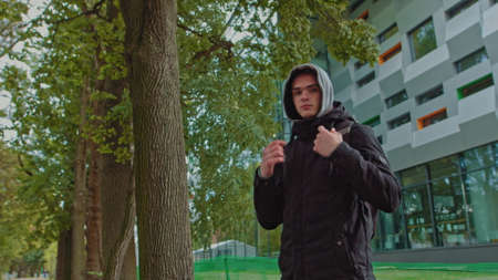 Portrait of a hipster young man putting on a hood while looking at the camera. Close up view of millennial boy in dark hoodie smiling standing on the street. People emotion conceptの写真素材
