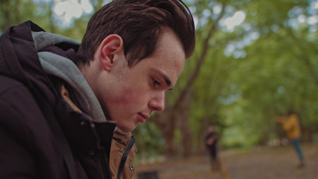 Young guy with a phone in his hands sitting on a bench in the park. In the background, a group of...