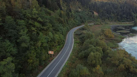 Aerial view of car driving through the forest and the lake on the side. Beautiful mountain road. Driving on the mountain road. Road trip. Mountain forest lake landscape. Car driving through pine forest.の写真素材
