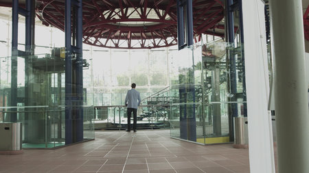 A stylishly dressed man walks down a glass corridor in a modern office. Shooting from the back of...