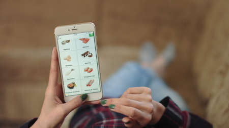 Woman at Home Lying on Couch in Living Room Using Smartphone Buys in Internet Shop. Woman Orders Seafood Home In An Online Store Using a Smartphone. Female Selects Seafood in Grocery Online Store. POVの写真素材