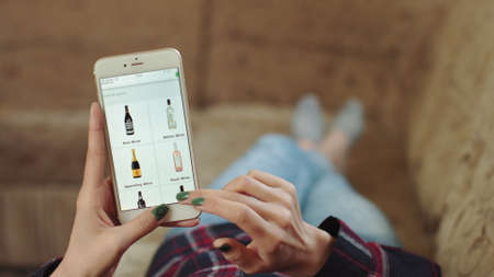 Woman at Home Lying on Couch in Living Room Using Smartphone Buys in Internet Shop. Woman Orders Wines Home In An Online Store Using a Smartphone. Female Selects Wines in Grocery Online Store. POVのeditorial素材