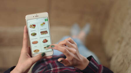 Woman at Home Lying on Couch in Living Room Using Smartphone Buys in Internet Shop. Woman Orders Prepared Food Home In An Online Store Using a Smartphone. Female Selects Prepared Food in Groceryの写真素材