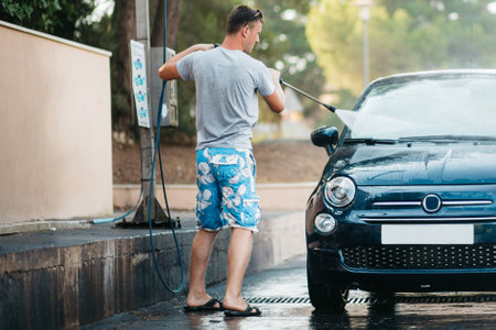 Young man washing a car with water under pressure in an outdoor sink. Manual washing with water under pressure in the sink on the street. Summer car wash.の写真素材