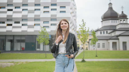 Student girl with backpack walking to the university building. Happy and smiling, looking at the camera and walking forward. Portrait of Female Student in White T-Shirt Confidently Looking into Cameraの写真素材