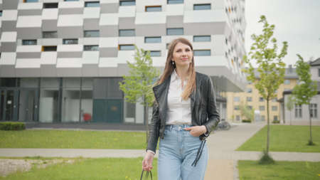 Portrait of Female Student in White T-Shirt Confidently Looking into Camera While Taking Walk in City. Happy Young Woman Enjoying Fine Sunny Day Outdoors. Slow Motion Backward Tracking Shot.の写真素材