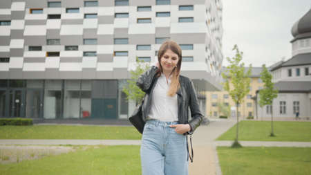 Portrait of Female Student in White T-Shirt Confidently Looking into Camera While Taking Walk in City. Happy Young Woman Enjoying Fine Sunny Day Outdoors. Slow Motion Backward Tracking Shot.の写真素材