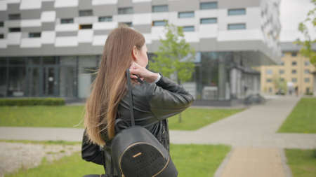 A portrait of a young girl, a student near a college or modern office, walking down the street with a backpack. Stylishly Dressed Modern Girl Slowly Puts a shoulder bag. Shooting from the backの写真素材