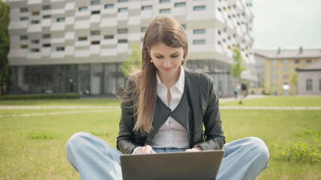 Happy woman working on laptop computer on green lawn in summer park. Young woman freelancer happy to receive new order. Female student using notebook outdoor. Teenage girl reading good news on laptopの写真素材