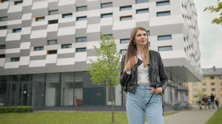 Stylishly dressed girl having fun outdoors in jeans clothes and leather jacket. Looks happy and pleasant. Look chic. Portrait of a female student on the background of the building. With nice Makeupの写真素材
