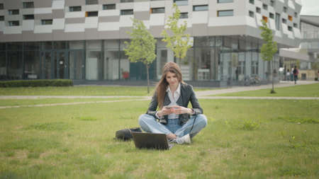 Happy young beautiful woman in stylish clothes, texting message on smartphone on city street. Portrait of an attractive female student sitting on the grass near school or college or officeの写真素材