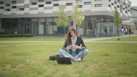 Happy young beautiful woman in stylish clothes, texting message on smartphone on city street. Portrait of an attractive female student sitting on the grass near school or college or officeの写真素材