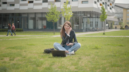 Teen woman, listening to music in headphone use smartphone at city walk sunset look around smile portrait, slow motion. Attractive female student sitting on the grass near school or college or officeの写真素材