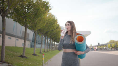 Young woman running drink water after workout outdoors in the city. Portrait close up of a young female, looking to camera. Rug In Her Hands And Water, Running Up The Stairs In The City Center.の写真素材