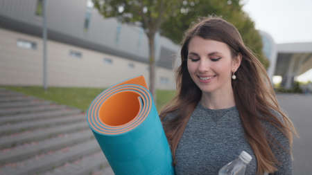 Portrait of a beautiful fitness girl. The Young Girl Goes Slowly To The Camera And Smiles. Active people. Inspiration and motivation. Active woman with fitness mat and water bottle in hands walkingの写真素材