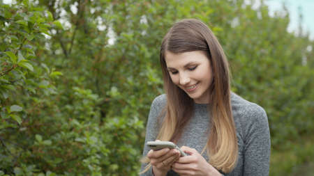 Happy young woman use smartphone standing in park sun outdoor health hand girl tree sport fitness summer nature internet technology female runner exercise phone workout mobile cell phoneの写真素材