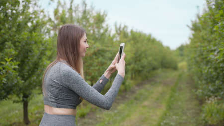 Photo on smartphone against the background of trees, beautiful girl photographing spring trees on smart phone. The average plan. Happy woman typing by mobile phone outdoors. Cheerful girl in parkの写真素材
