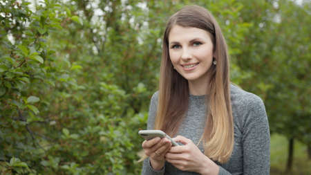 Happy young woman use smartphone standing in park sun outdoor health hand girl tree sport fitness summer nature internet technology female runner exercise phone workout mobile cell phoneの写真素材