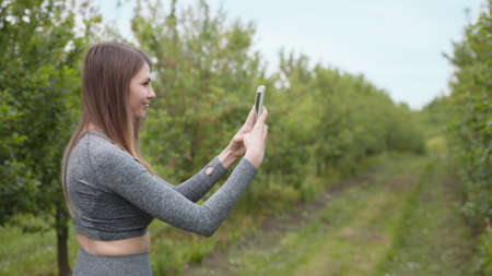 Photo on smartphone against the background of trees, beautiful girl photographing spring trees on smart phone. The average plan. Happy woman typing by mobile phone outdoors. Cheerful girl in parkの写真素材