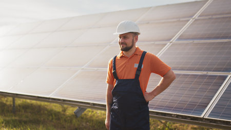 Close up portrait of male electrical worker in protective helmet standing near solar panel. Clean energy production. Green energy. Solar farm. Cheerful man smiling while standing at solar power farmの写真素材