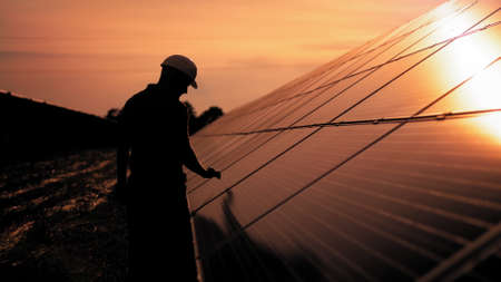 Assistance technical worker in uniform is checking an operation and efficiency performance of photovoltaic solar panels. Unidentified solar power engineer touches solar panels with his hand at sunsetの写真素材