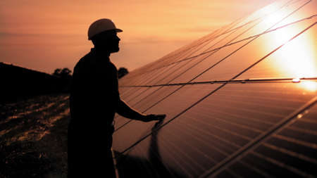 Assistance technical worker in uniform is checking an operation and efficiency performance of photovoltaic solar panels. Unidentified solar power engineer touches solar panels with his hand at sunsetの写真素材