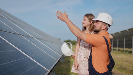 A father, a solar power engineer, and his daughter are standing near solar panels. The father explains to the child the principle of solar electricity and puts a protective helmet on the girls head.の写真素材