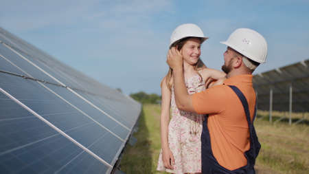 A father, a solar power engineer, and his daughter are standing near solar panels. The father explains to the child the principle of solar electricity and puts a protective helmet on the girls head.の写真素材