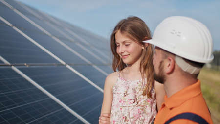 Father with little girl at solar power plant. The father talks about solar energy. The concept of green energy will save the planet for children. The father puts a protective helmet on the girls headの写真素材