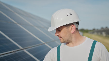 Portrait of happy male engineer in protective helmet looking to camera. Handsome man in uniform smiling while standing at solar power farm. Concept of green energy. Ecological future.の写真素材