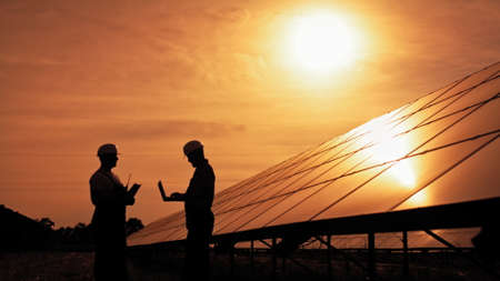 Two unidentified solar power engineers talking to each other at sunset. Couple of workers examining solar batteries and communicating on business. Ecological future. Sustainable energy.の写真素材