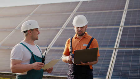 Technician with laptop checking the panels at solar energy installation. Business cooperation. Solar park. Alternative energy concept. Clean energy. Two workers in a uniform and hard hatの写真素材