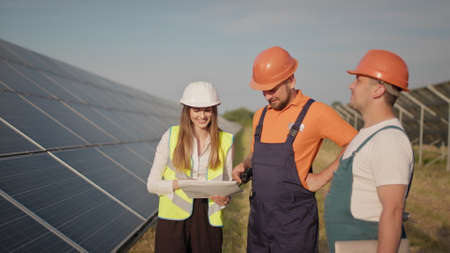 Engineers use drawings on the solar farm in special uniform standing look around discuss the installation of sunny batteries. Successful business. Ecological construction. Solar panels field. Teamworkの写真素材